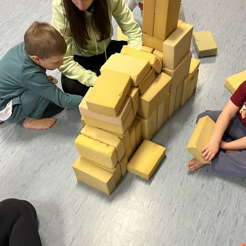 Kinder und eine Erwachsene bauen aus gelben Baukl&ouml;tzen eine Struktur auf einem blauen Boden.
