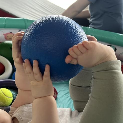 Ein Baby hält einen blauen Ball mit beiden Händen, seine Füße sind in einer bunten Spielumgebung sichtbar.