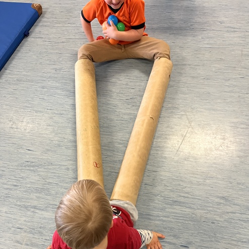 Zwei Kinder spielen auf dem Boden, eines sitzt in einem Holzrohr, das als „V“ geformt ist, während das andere steht.