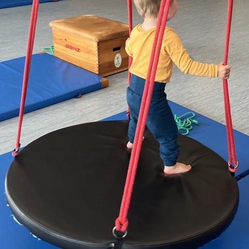 Ein Kleinkind steht barfuß auf einer runden, schwingenden Matte in einem Spielraum mit blauen Matten.