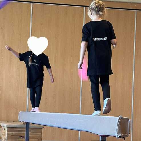 Zwei Kinder auf einem Schwebebalken in einer Turnhalle, eines mit einem Lappen, das andere in schwarzem T-Shirt.