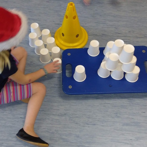 Kind mit Weihnachtsmütze sitzt auf dem Boden und stapelt weiße Becher neben einem blauen Tablett und einem gelben Pylon.
