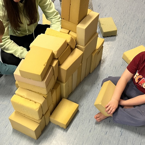 Kinder und eine Erwachsene bauen aus gelben Bauklötzen eine Struktur auf einem blauen Boden.