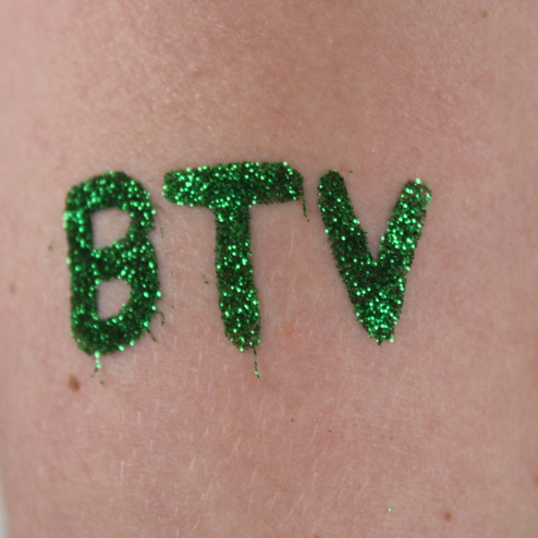 Gr&uuml;ne Glitzerbuchstaben &bdquo;BTV&ldquo; auf einem Arm, vor einem unscharfen Hintergrund.