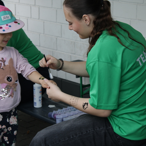 Kind mit buntem Cap erh&auml;lt ein tempor&auml;res Tattoo von einer Person in gr&uuml;nem Shirt, w&auml;hrend sie auf einer Bank sitzen.