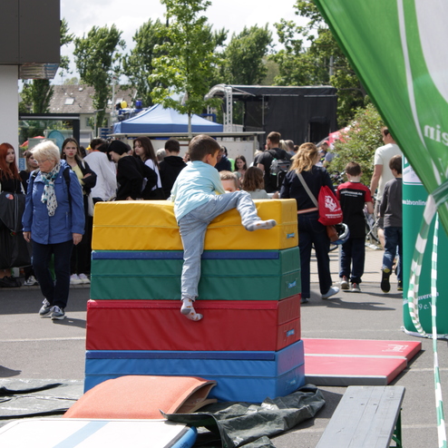 Ein Kind klettert auf einen bunten Schaumstoffblockstapel, w&auml;hrend Besucher eines Festes im Hintergrund laufen.