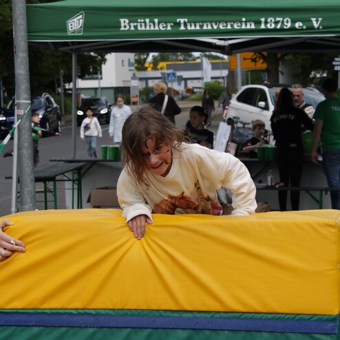 M&auml;dchen mit Gesichtsbemalung klettert &uuml;ber eine weiche, bunten Mattenbarriere vor einem Zelt des Br&uuml;hler Turnvereins.