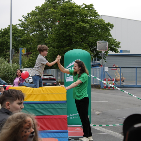 Kind auf einer bunten Spielmatte hoch oben, w&auml;hrend ein Betreuer ihm die Hand reicht; Zuschauer im Hintergrund.