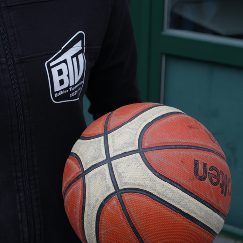 Nahaufnahme eines Basketballs, gehalten von einer Person in einem schwarzen Sweatshirt mit Logo.