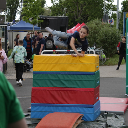 Kind springt &uuml;ber einen bunten Turnblock auf einer Sportveranstaltung, Zuschauer im Hintergrund.