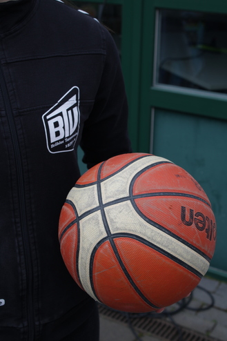 Nahaufnahme eines Basketballs, gehalten von einer Person in einem schwarzen Sweatshirt mit Logo.