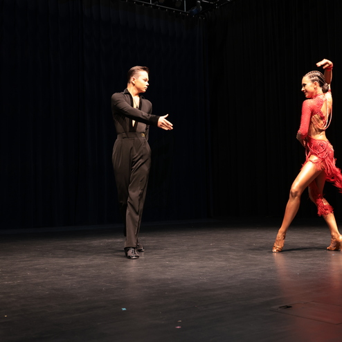 Tanzende Paare auf einer B&uuml;hne, der Mann in schwarzem Outfit, die Frau in einem roten, fransigen Kleid.