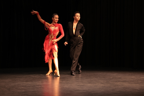 Tanzpaar in farbenfrohen Kostümen führt einen lebhaften Tanz auf einer Bühne mit schwarzem Hintergrund auf.