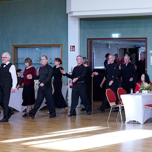 Tanzende Gruppe älterer Menschen in eleganter Kleidung beim Betreten eines festlich dekorierten Raums.