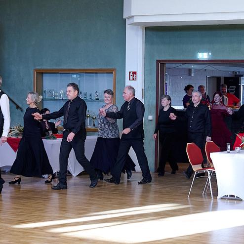 Tanzende Personen in eleganter Kleidung in einem festlichen Saal mit Tischen im Hintergrund.