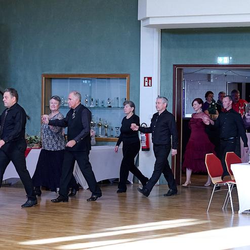 Tanzende Paare in eleganter Kleidung betreten einen Saal, Zuschauer sitzen an Tischen im Hintergrund.