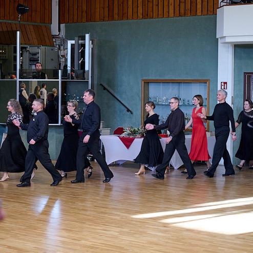 Tanzende Paare in eleganter Kleidung gehen in einer Festhalle aufeinander zu. Tische sind im Hintergrund sichtbar.
