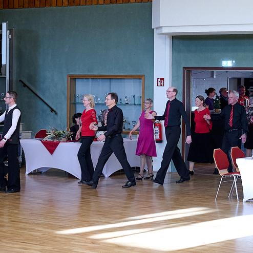 Tanzende Paare in formeller Kleidung bewegen sich durch einen festlich dekorierten Festraum.