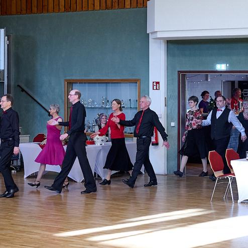 Tanzende Paare in eleganter Kleidung bewegen sich in einem festlichen Saal mit Tischen im Hintergrund.