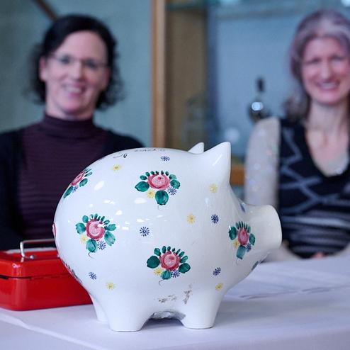 Dekoratives Sparschwein mit Blumenmustern auf einem Tisch, im Hintergrund zwei Frauen sitzen.
