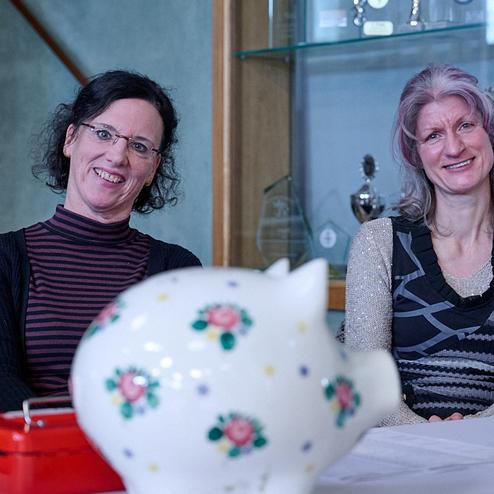 Zwei Frauen lächeln am Tisch, ein buntes Sparschwein und eine rote Box sind sichtbar im Vordergrund.