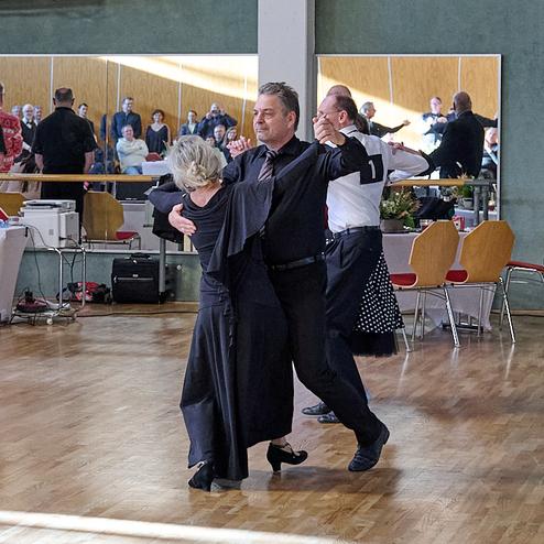 Zwei Paare tanzen in eleganter Kleidung auf einer Tanzfläche, im Hintergrund Zuschauer und festliche Dekoration.