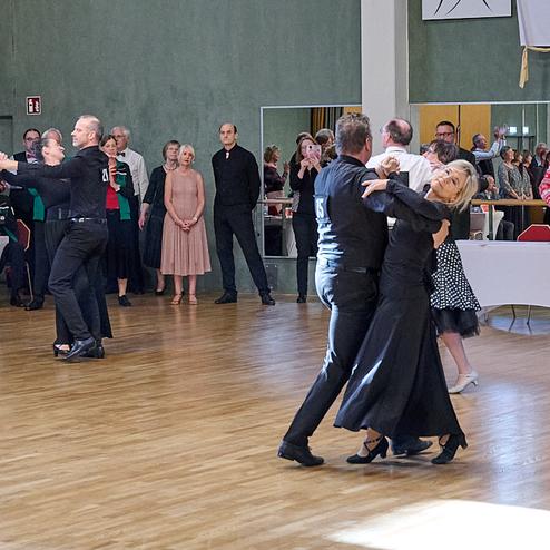 Tanzende Paare in eleganter Kleidung auf einer Tanzfläche, umgeben von applaudierenden Zuschauern bei einer Veranstaltung.