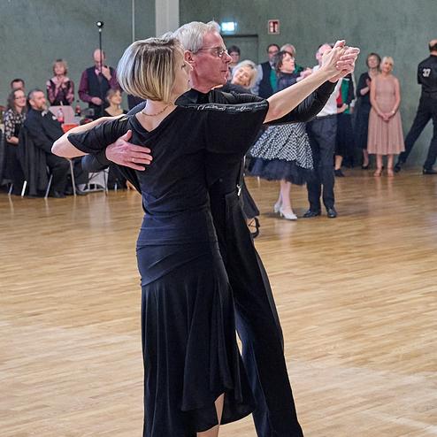 Tanzendes Paar in eleganter Kleidung auf einem Parkettboden, im Hintergrund Zuschauer bei einer Tanzveranstaltung.