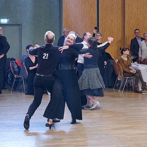 Tanzpaar in eleganter Kleidung auf einer Veranstaltung, umgeben von Zuschauern, die applaudieren und zuschauen.