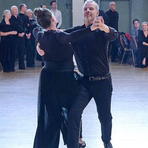 Tanzpaar in eleganter Kleidung führt einen Tanz auf einer Halle mit Zuschauern im Hintergrund aus.