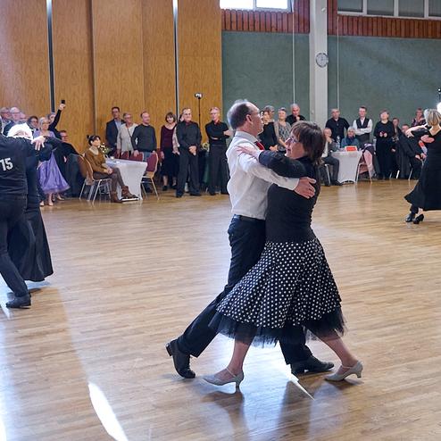 Tanzpaar im Halbprofil auf einer Tanzfläche, umgeben von Zuschauern in festlicher Kleidung.