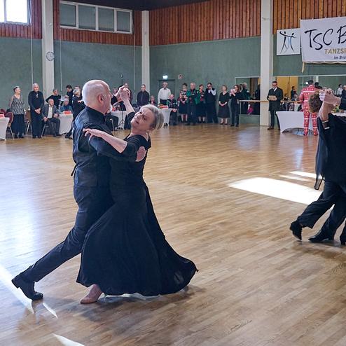 Tanzpaare in eleganter Kleidung tanzen auf einer hellen Holzfläche während einer Tanzveranstaltung im Hintergrund.