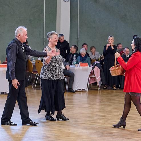 Tanzpaar auf einer Veranstaltung, während eine Frau in rotem Mantel einen Korb hält, Zuschauer im Hintergrund.