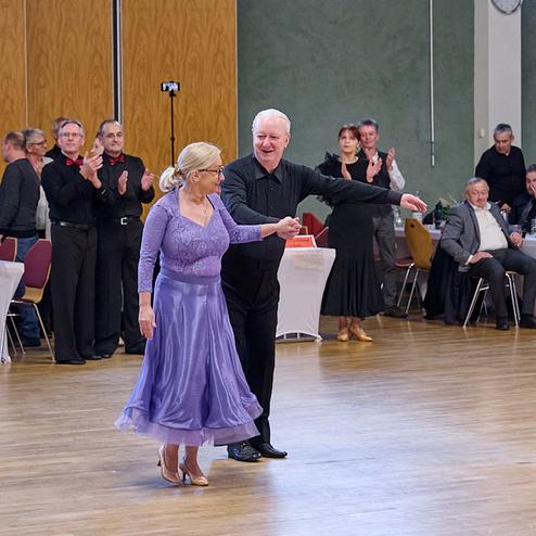 Tanzpaar in eleganter Kleidung bei einem Tanzabend, umgeben von applaudierendem Publikum in einem festlichen Saal.