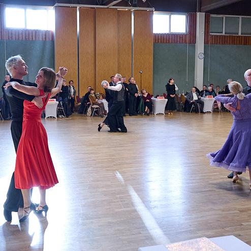 Tanzveranstaltung in einem Saal mit Paaren in eleganter Tanzkleidung auf Holzfußboden.