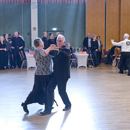 Tanzendes Paar in eleganter Kleidung auf einer Tanzfläche, umgeben von Zuschauern in einem festlichen Ambiente.