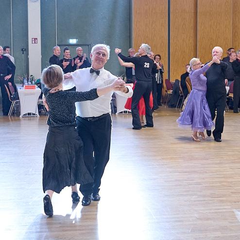 Tanzpaare präsentieren sich auf einer Fläche, umgeben von Zuschauern in einem festlichen Saal.