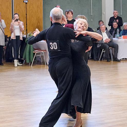 Tanzendes Paar in eleganter Kleidung auf einer Tanzfläche, umgeben von Zuschauern in einem Festsaal.