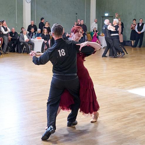 Tanzpaar in Bewegung auf einer Tanzfläche, umgeben von Zuschauern in einem festlichen Saal.