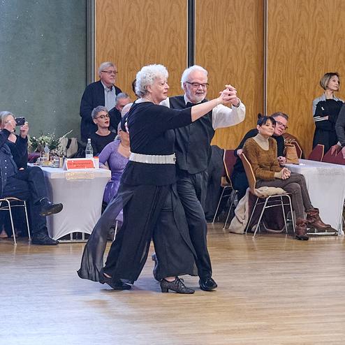 Tanzendes Paar in eleganter Kleidung auf einer Tanzfläche, Zuschauer beobachten in einem festlichen Saal.