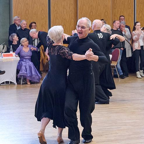 Tanzendes Paar im Schwarzen, umgeben von Zuschauern in einem festlichen Saal. Holzfußboden und große Fenster im Hintergrund.