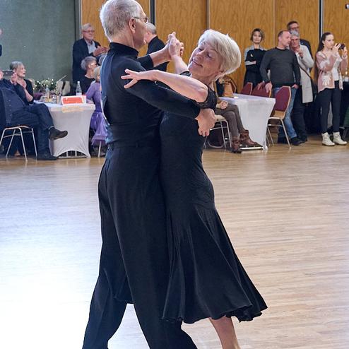 Tanzendes Paar in eleganter Kleidung auf einer Tanzfläche, umgeben von Zuschauern in einem festlichen Saal.