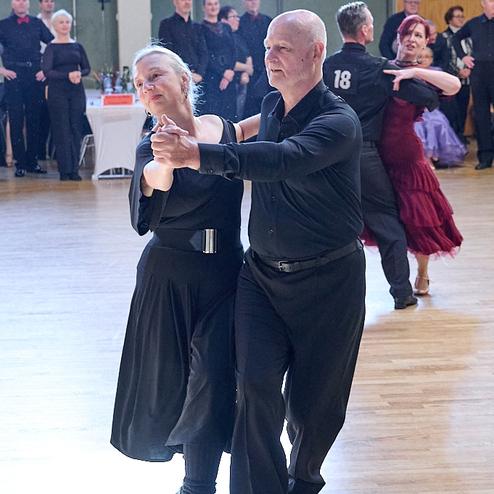 Tanzendes Paar in eleganter Kleidung auf einer Tanzfläche, umgeben von weiteren Tänzern und Zuschauern.