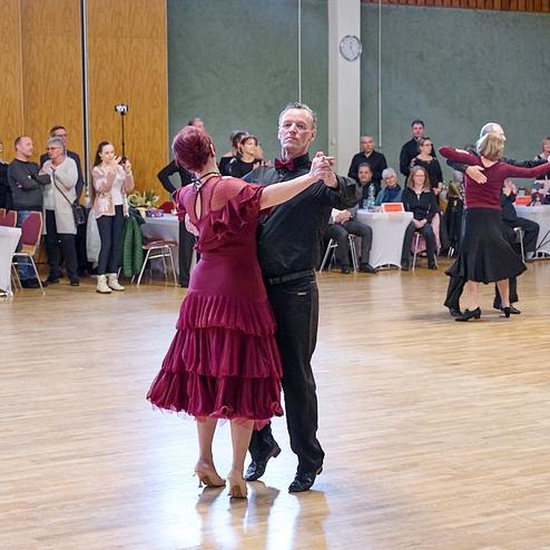 Tanzpaar in eleganter Kleidung tanzt in einem Saal, umgeben von zuschauenden Personen. Holzfußboden und Dekor sichtbar.