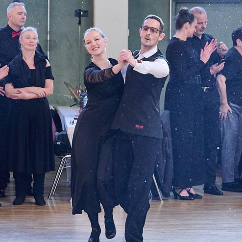 Tanzendes Paar in eleganter Kleidung auf einer Tanzfläche, umgeben von applaudierenden Zuschauern.