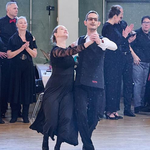 Tanzpaar in eleganter Kleidung führt einen Schritt auf der Tanzfläche, umgeben von applaudierenden Zuschauern.