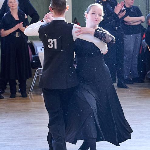 Tanzpaar in eleganter Kleidung tanzt im Fokus auf einer Tanzfläche, Zuschauer applaudieren im Hintergrund.