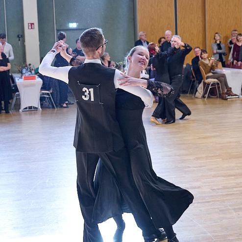 Tanzpaar in festlicher Kleidung tanzt auf einer Tanzfläche, Zuschauer applaudieren im Hintergrund.
