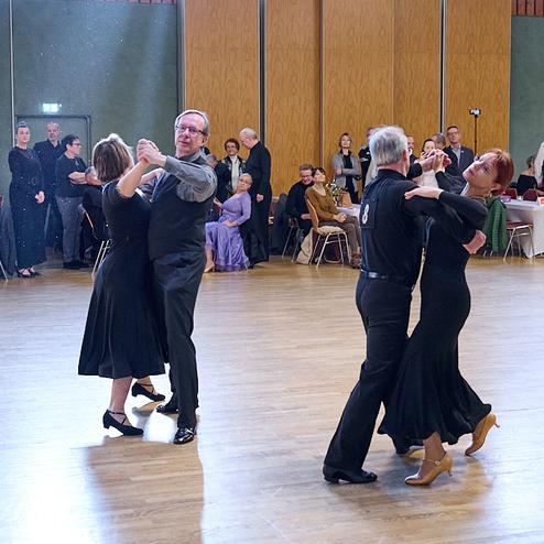Tanzpaare in eleganter Kleidung tanzen auf einer Holzfläche während einer Tanzveranstaltung, im Hintergrund Zuschauer.