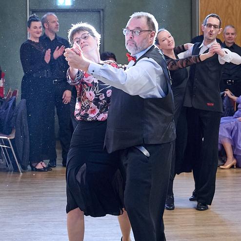 Tanzpaar in eleganter Kleidung führt einen Walzer auf einer Tanzfläche, Zuschauer applaudieren im Hintergrund.